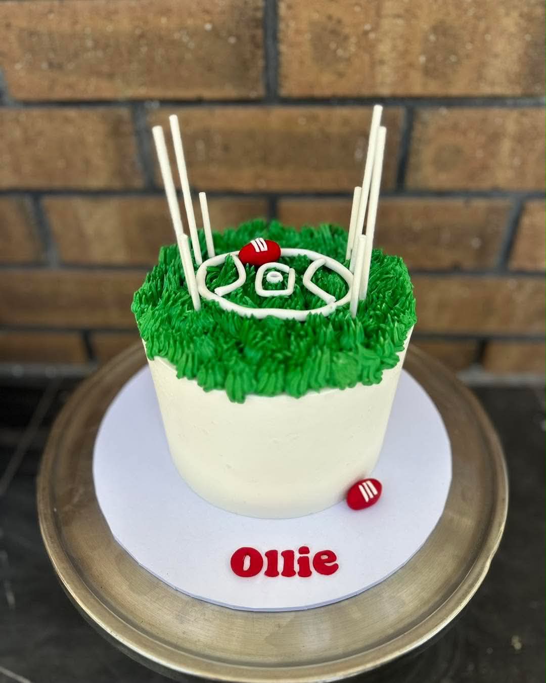 White and green AFL football birthday cake for Ollie with green grass buttercream top, white goal posts, red AFL ball and Ollie name plaque